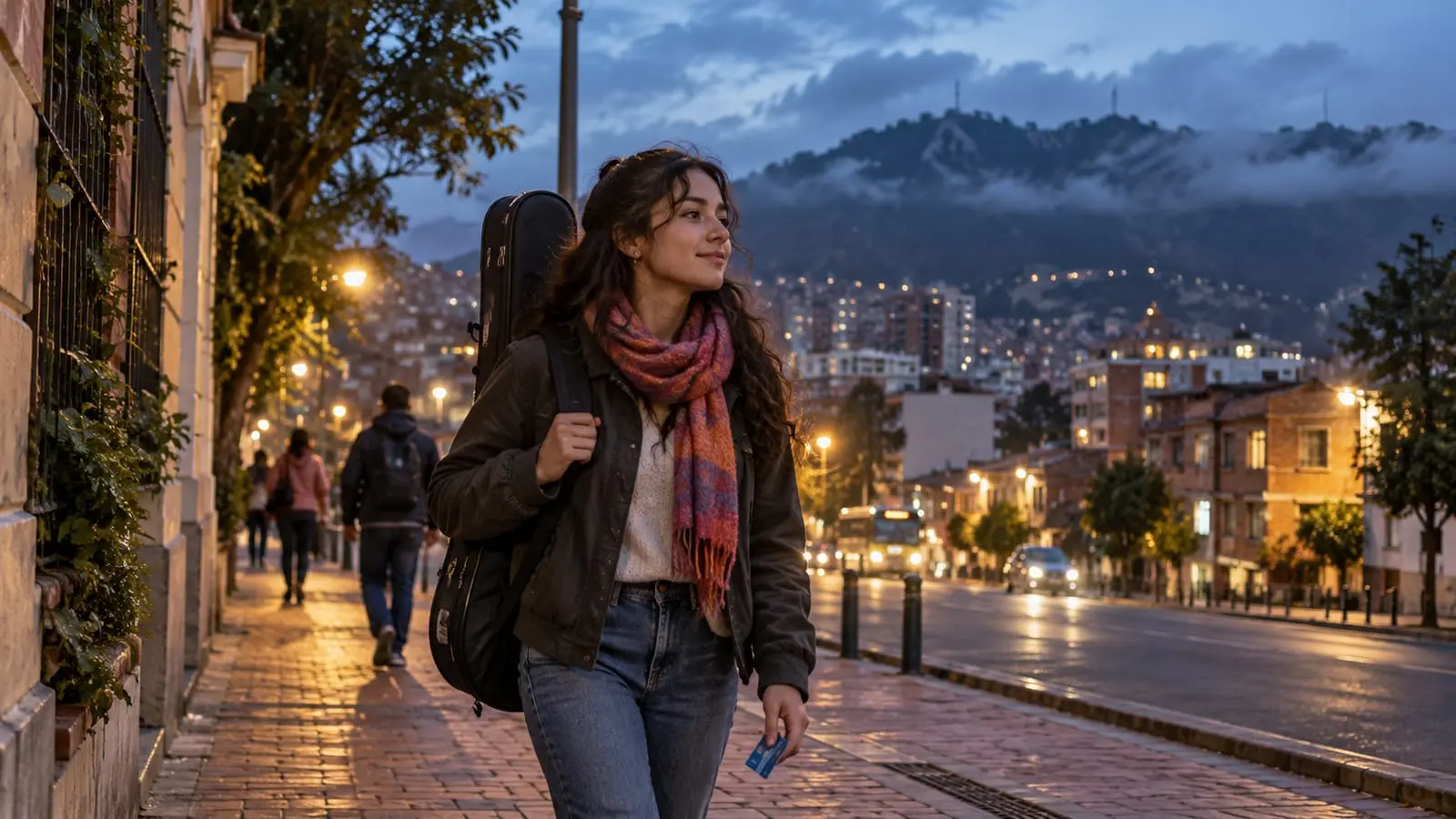 A young woman with a guitar case over her shoulder walks down a Bogotá street at sunset, with the mountains in the background